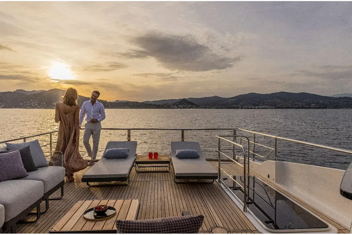 A couple on a yacht at sunset, enjoying the view over the water, with loungers and a table of fruit nearby.
